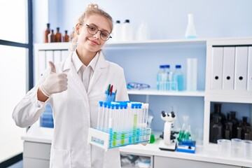 Young caucasian woman working at scientist laboratory holding samples smiling happy and positive, thumb up doing excellent and approval sign