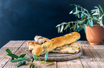 Group of bread sticks stuffed with green olives ion cutting board, olive tree branch, olives in bowl on wooden table with dark background