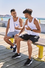 Two hispanic men sporty couple smiling confident sitting on bench at seaside