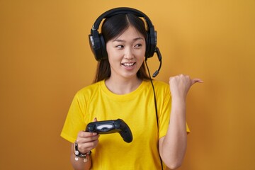 Chinese young woman playing video game holding controller smiling with happy face looking and pointing to the side with thumb up.