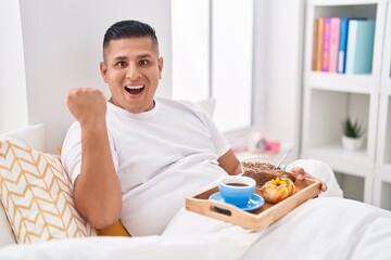Young hispanic man eating breakfast in the bed screaming proud, celebrating victory and success very excited with raised arms