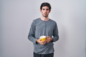 Young hispanic man wearing pajama drinking a cup of coffee skeptic and nervous, frowning upset...