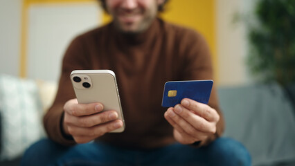 Young hispanic man using smartphone and credit card sitting on sofa at home