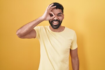 Fototapeta na wymiar Hispanic man with beard standing over yellow background doing ok gesture with hand smiling, eye looking through fingers with happy face.