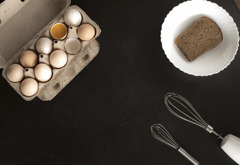 Eggs, brown piece of bread and whisk for whipping on black background