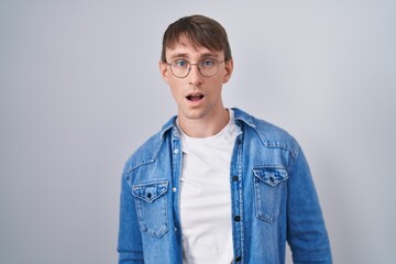 Caucasian blond man standing wearing glasses in shock face, looking skeptical and sarcastic, surprised with open mouth