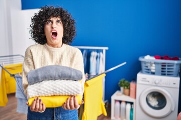 Young brunette woman with curly hair holding clean laundry angry and mad screaming frustrated and...