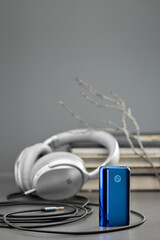 Headphones with a wire against the background of books. In the foreground is a blue electronic tobacco heating device, studio shot.