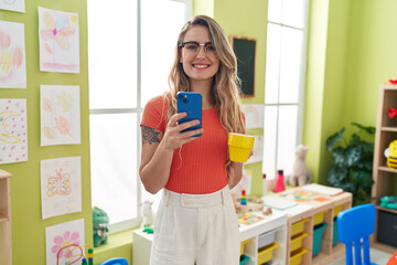 Young blonde woman teacher using smartphone drinking coffee at kindergarten