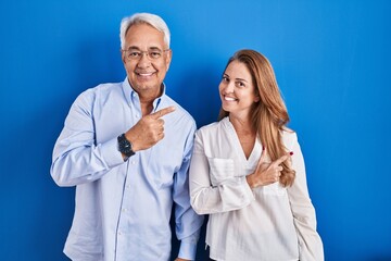Middle age hispanic couple standing over blue background cheerful with a smile on face pointing with hand and finger up to the side with happy and natural expression