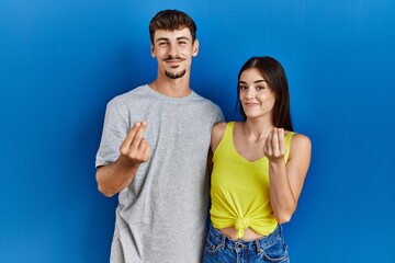 Young hispanic couple standing together over blue background doing money gesture with hands, asking for salary payment, millionaire business
