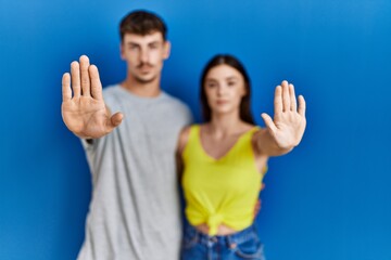 Young hispanic couple standing together over blue background doing stop sing with palm of the hand. warning expression with negative and serious gesture on the face. - Powered by Adobe