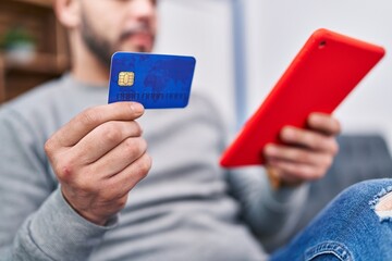 Young latin man using touchpad and credit card sitting on sofa at home
