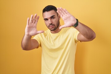 Young hispanic man standing over yellow background doing frame using hands palms and fingers, camera perspective