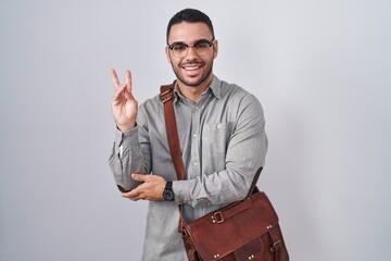 Young hispanic man wearing suitcase smiling with happy face winking at the camera doing victory...