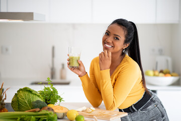 Smiling young african american female vegan at table with organic vegetables hold glass with fresh...