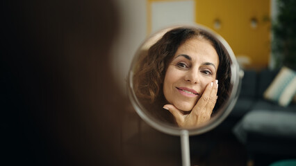 Middle age hispanic woman sitting on sofa looking face on mirror at home