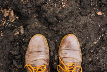 The worker is standing in two brown leather boots with long laces against the background of the earth, black soil, soil. Background, texture, farming, agriculture.
