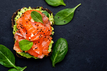 Avocado salmon sandwich or toast on rye bread with spinach, crushed cashew nuts and sesame seeds, black table background, top view