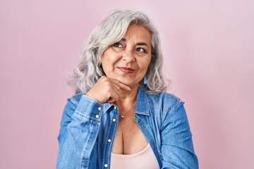 Middle age woman with grey hair standing over pink background with hand on chin thinking about...