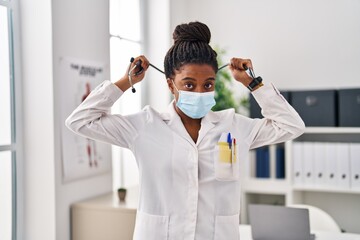 African american woman wearing doctor uniform and medical mask holding stethoscope at clinic