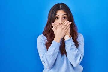 Hispanic young woman standing over blue background shocked covering mouth with hands for mistake. secret concept.