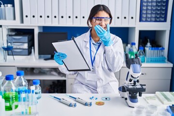 Hispanic young woman working at scientist laboratory laughing and embarrassed giggle covering mouth...