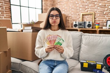 Young hispanic woman sitting on the sofa at new home holding money puffing cheeks with funny face....