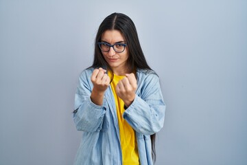 Young hispanic woman standing over blue background ready to fight with fist defense gesture, angry...