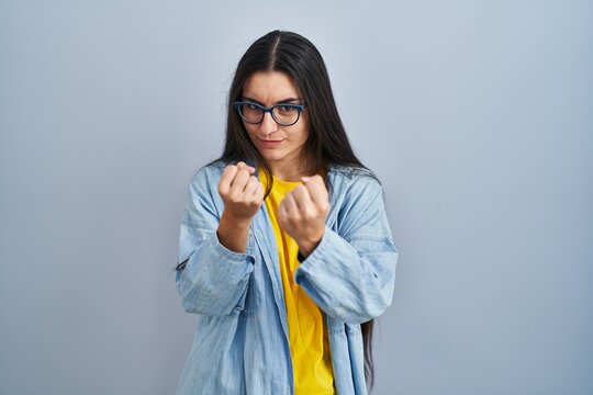 Young hispanic woman standing over blue background ready to fight with fist defense gesture, angry and upset face, afraid of problem