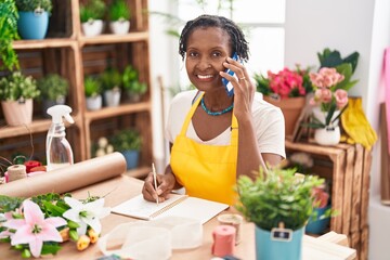 Middle age african american woman florist talking on smartphone writing on notebook at flower shop