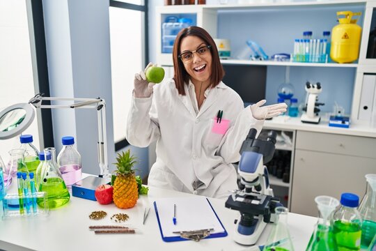 Young hispanic woman working at scientist laboratory holding fruit celebrating achievement with happy smile and winner expression with raised hand
