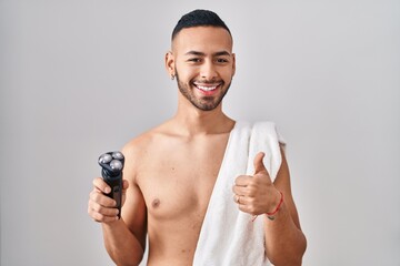 Young hispanic man holding electric razor smiling happy and positive, thumb up doing excellent and approval sign - obrazy, fototapety, plakaty