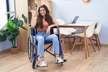 Fototapeta premium Young teenager girl sitting on wheelchair at the living room pointing thumb up to the side smiling happy with open mouth