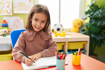 Adorable hispanic girl preschool student sitting on table writing on notebook at kindergarten