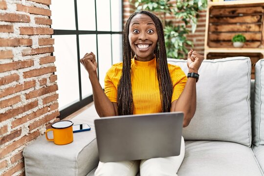 African woman sitting on the sofa using laptop at home screaming proud, celebrating victory and success very excited with raised arms