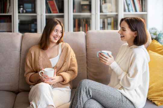 Laughing european millennial ladies in sweaters with coffee cups talk, sit on sofa, enjoy visit, free time