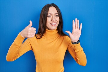 Young brunette woman standing over blue background showing and pointing up with fingers number six while smiling confident and happy.