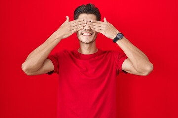 Young hispanic man standing over red background covering eyes with hands smiling cheerful and funny. blind concept.