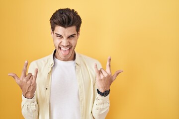 Young hispanic man standing over yellow background shouting with crazy expression doing rock symbol...