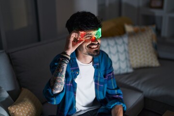 Young hispanic man using augmented reality glasses sitting on sofa at home
