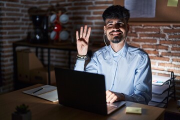 Young hispanic man with beard working at the office at night showing and pointing up with fingers number four while smiling confident and happy.