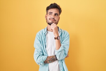 Young hispanic man with tattoos standing over yellow background with hand on chin thinking about question, pensive expression. smiling and thoughtful face. doubt concept.