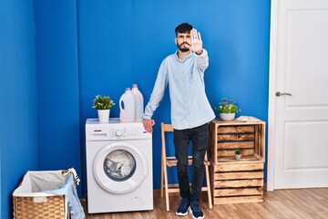 Young hispanic man with beard doing laundry standing at laundry room with open hand doing stop sign with serious and confident expression, defense gesture