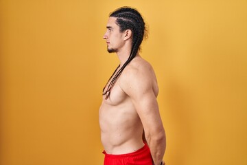 Fototapeta premium Hispanic man with long hair standing shirtless over yellow background looking to side, relax profile pose with natural face and confident smile.