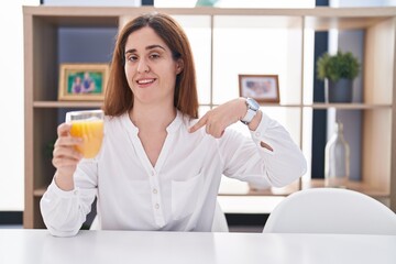 Brunette woman drinking glass of orange juice looking confident with smile on face, pointing oneself with fingers proud and happy.