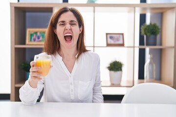 Brunette woman drinking glass of orange juice angry and mad screaming frustrated and furious, shouting with anger. rage and aggressive concept.