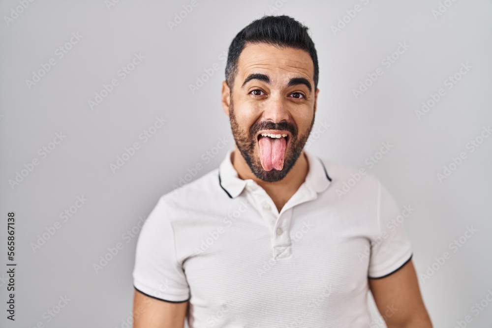 Sticker young hispanic man with beard wearing casual clothes over white background sticking tongue out happy