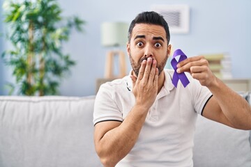 Young hispanic man with beard holding purple ribbon awareness covering mouth with hand, shocked and...