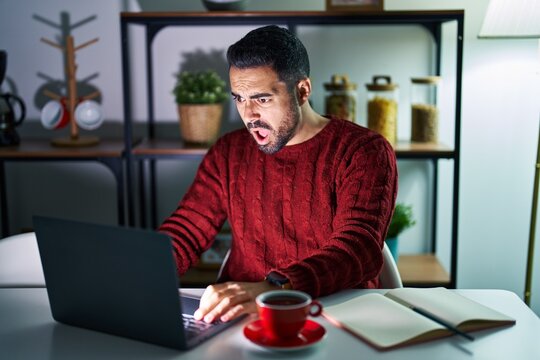 Young Hispanic Man With Beard Using Computer Laptop At Night At Home In Shock Face, Looking Skeptical And Sarcastic, Surprised With Open Mouth